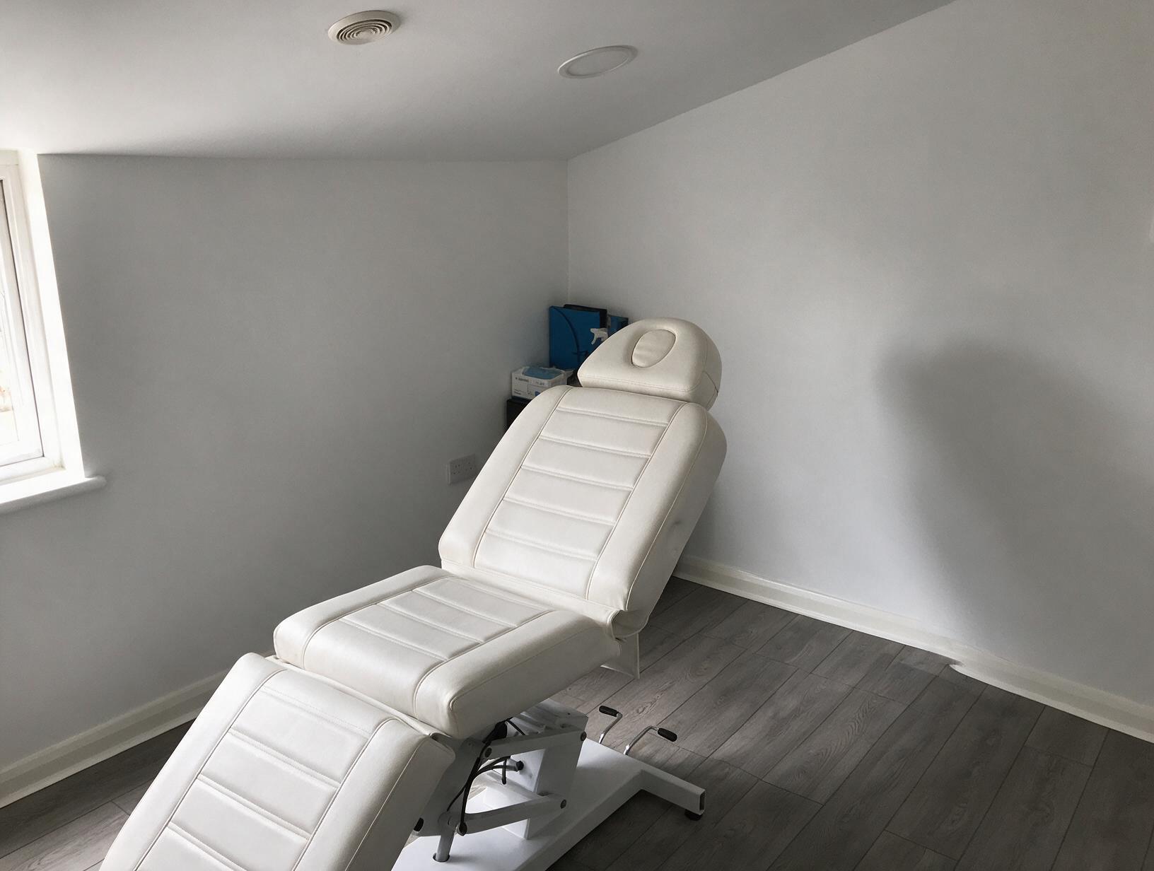 Empty treatment room with a white padded reclining couch, grey wood-effect floor, white walls and a window on the left.