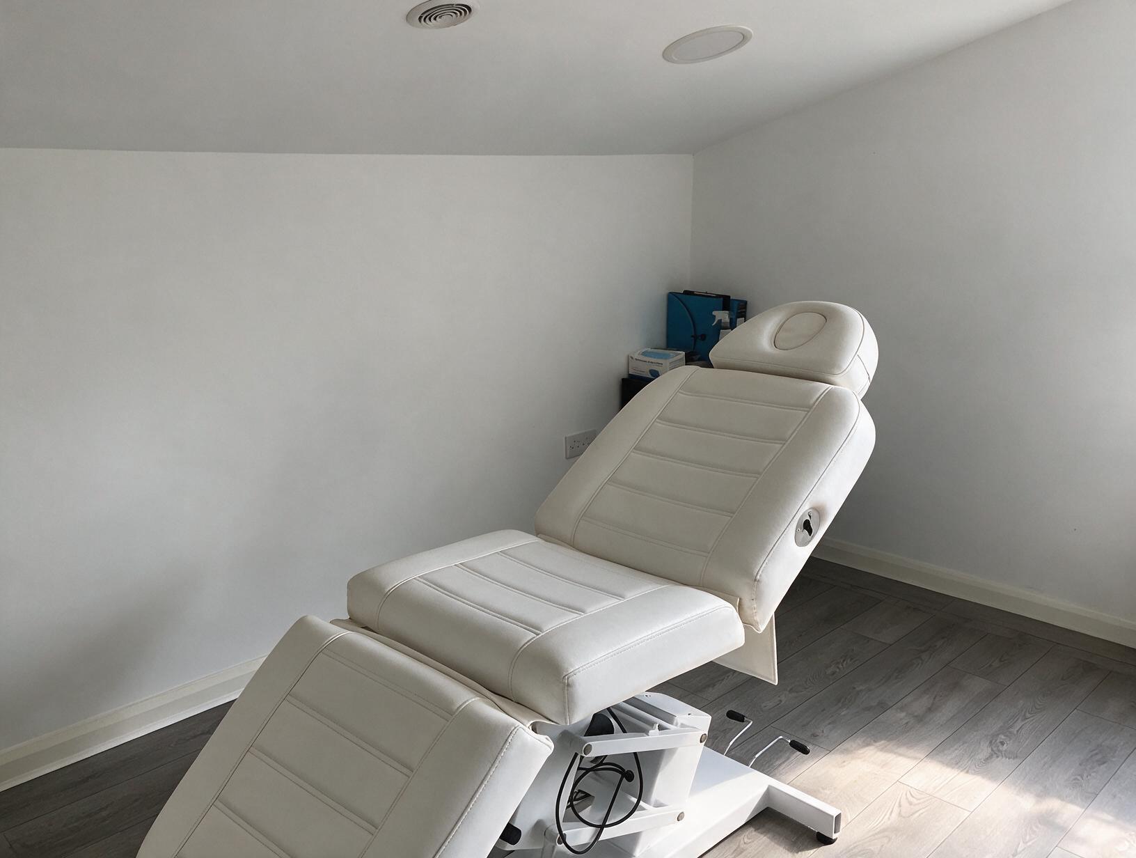 Empty treatment room with a white padded treatment couch, white walls, grey wood-effect floor and daylight on the floor.