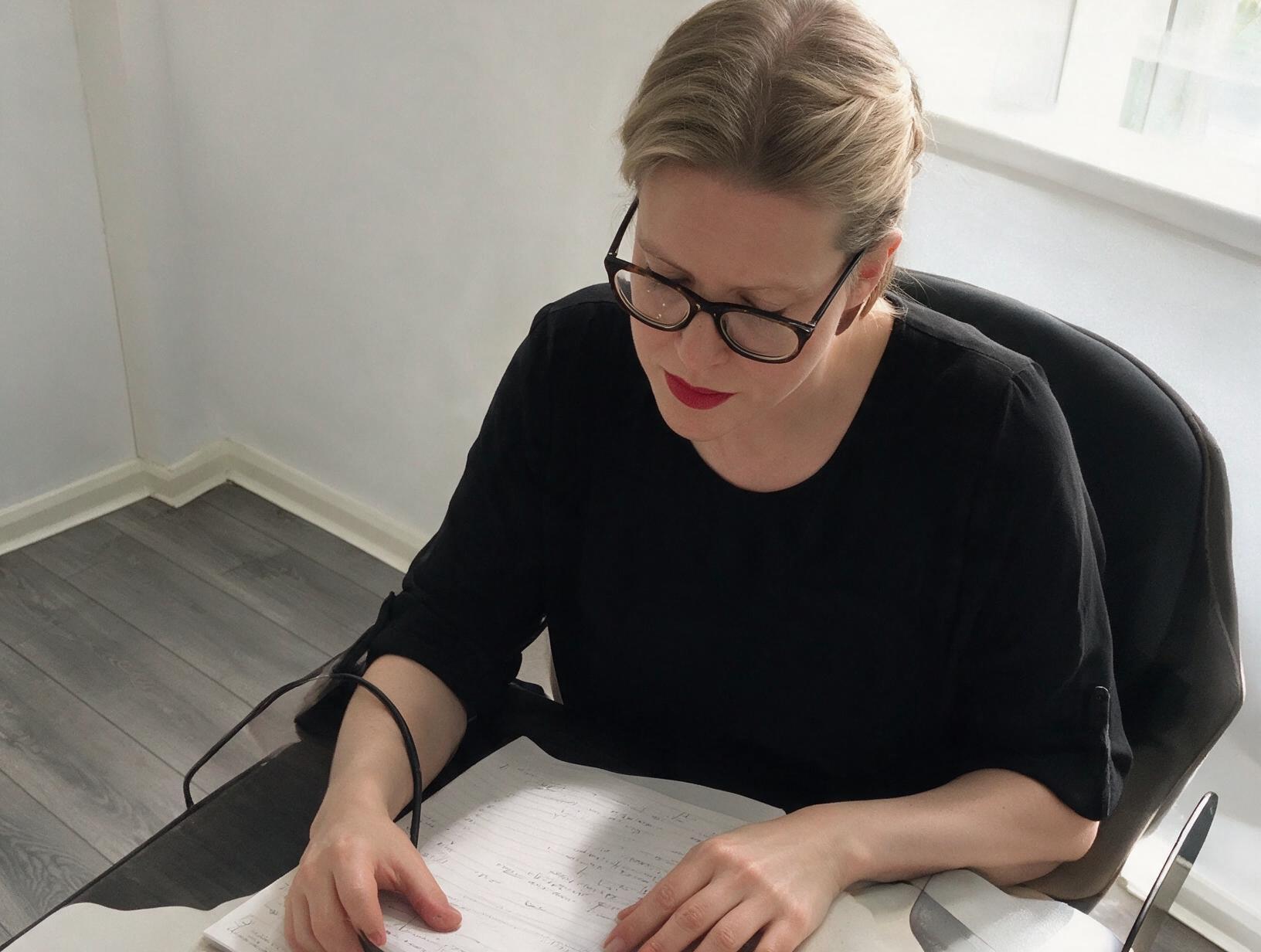 Woman with glasses, red lipstick and hair tied back, in a black top, looking down at handwritten notes at a desk by a window.